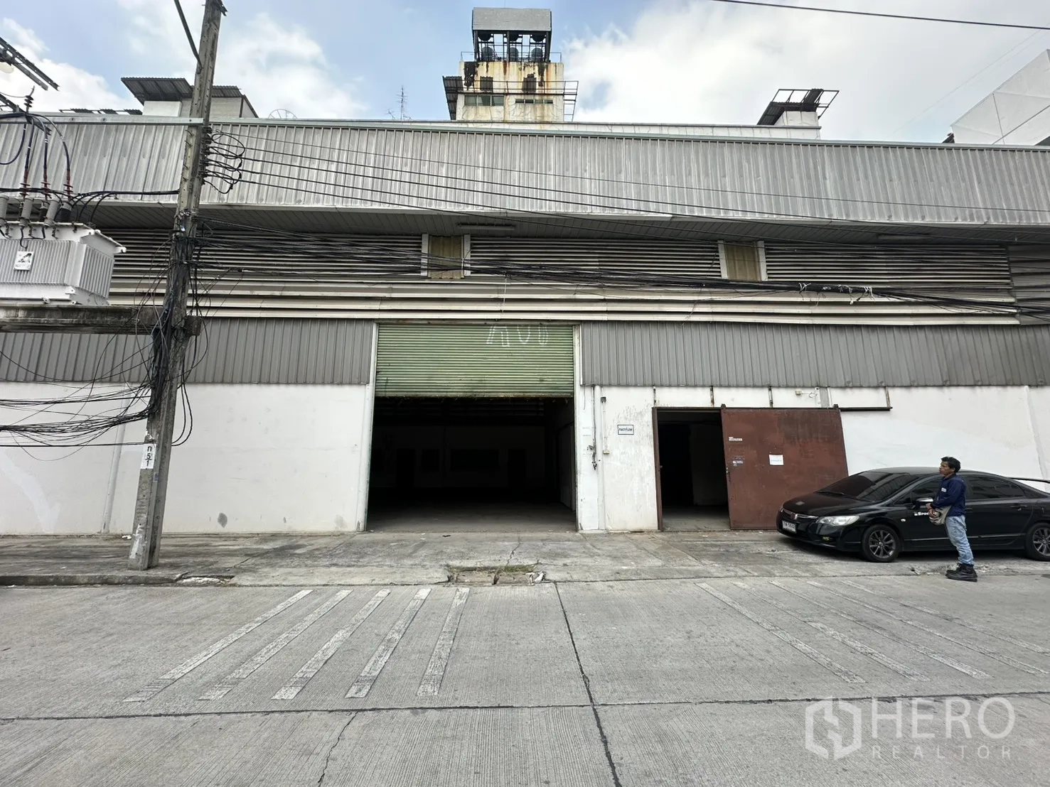 Warehouse for rent in Suan Luang, Bangkok - Front elevation of a metal-clad warehouse featuring a wide rolling door and parking space at the entrance.