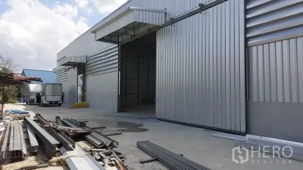 Warehouse for rent in Khlong Sam Wa, Bangkok - Side view of the warehouse loading area with open bay doors, parked trucks and steel materials on the driveway.
