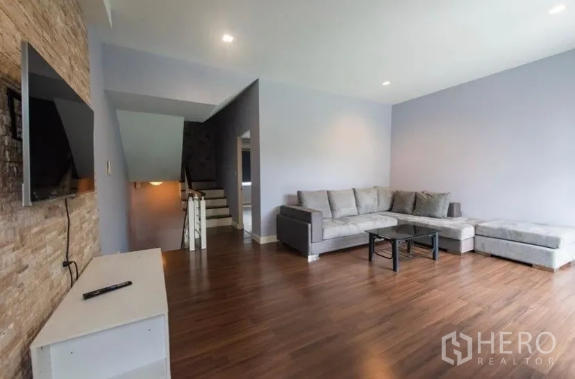 Townhouse for rent in Suan Luang, Bangkok - Spacious upstairs family room with wood flooring, grey L-shaped sofa and TV on a stone accent wall.