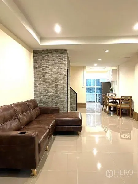 Townhouse for rent in Bang Na, Bangkok - Open-plan living room with brown leather sectional sofa and recessed ceiling lights.