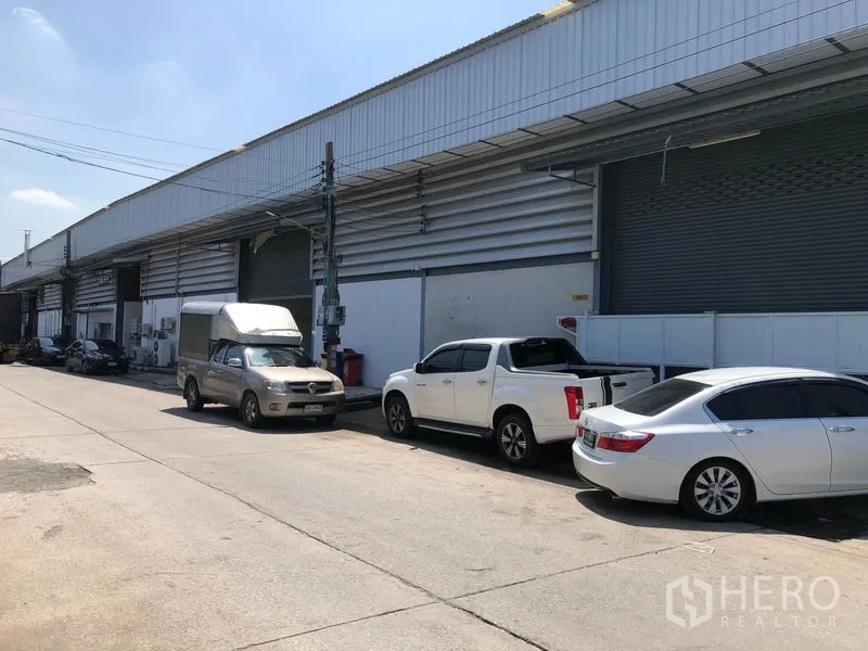 Warehouse for rent in Mueang Samut Prakan, Samut Prakan - Row of warehouses along a wide street with multiple roller shutters and roadside parking.