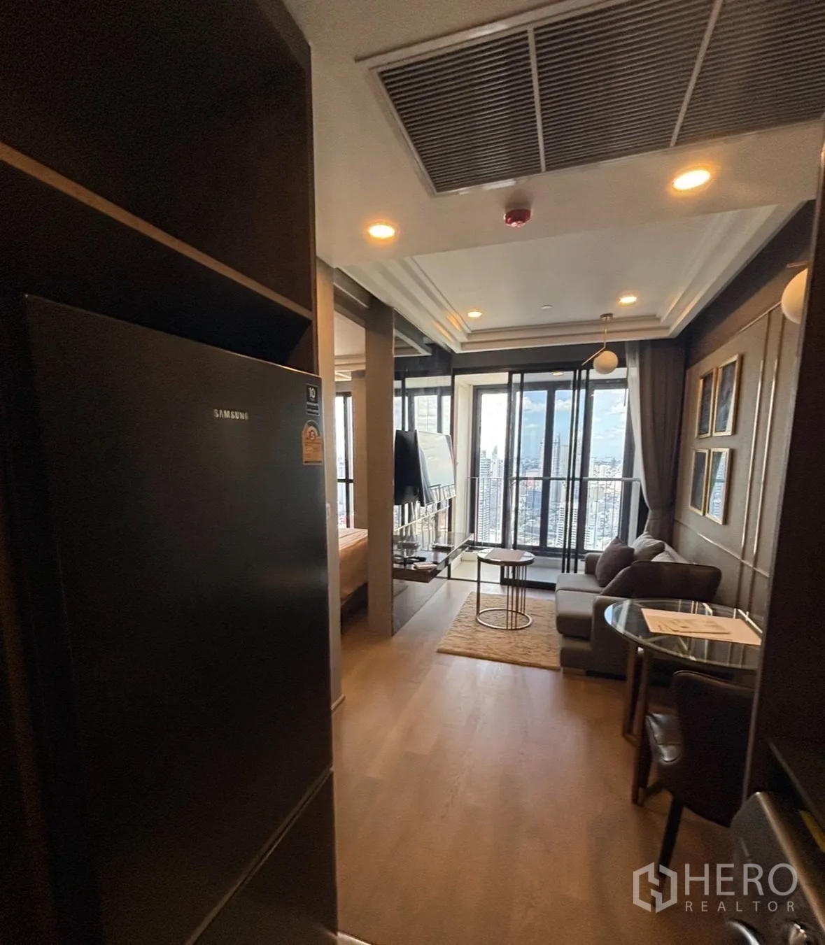 Condominium for rent in Bang Rak, Bangkok - Entry to an open-plan living area with sofa, dining set, TV, tall glass doors to a balcony, and a refrigerator.
