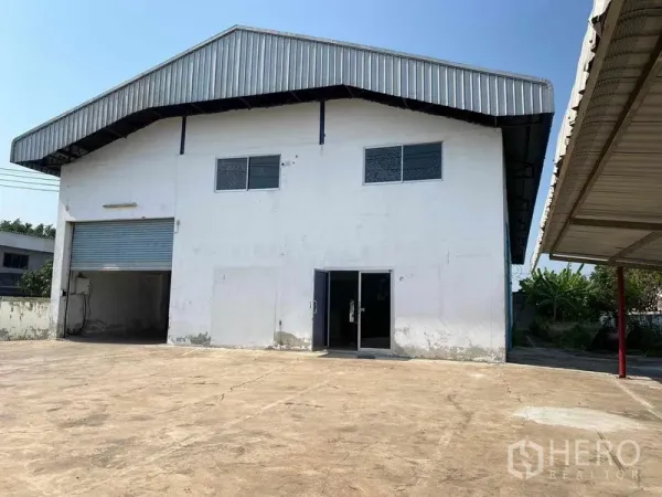 Factory for rent or sale in Bang Phli, Samut Prakan - Front exterior of a two-storey factory with metal roof, roller shutter, glass office door and a wide concrete yard.