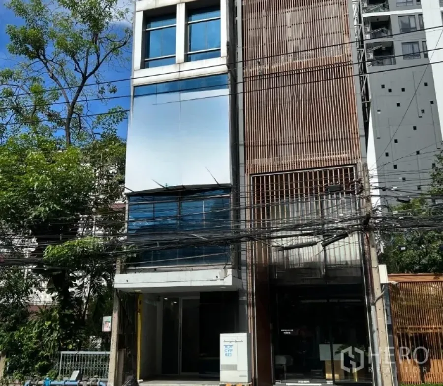 Retail Space for rent in Watthana, Bangkok - Four‑storey shophouse facade with glass panels and vertical wooden slats, trees and street cables in front.