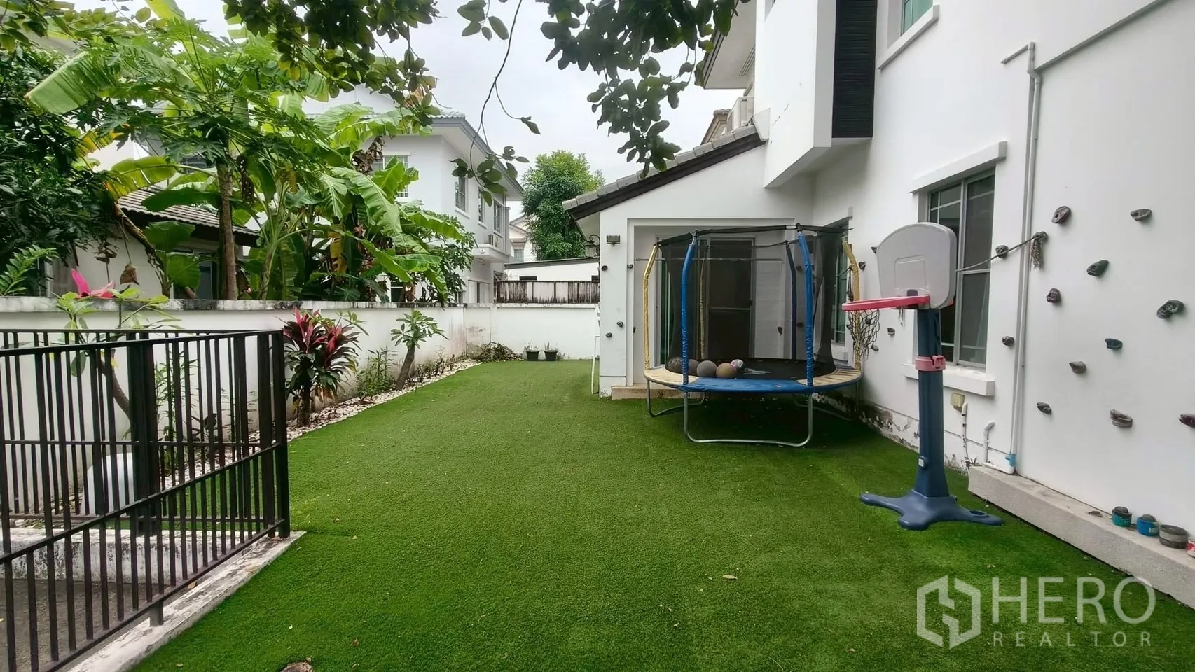 Detached House for rent or sale in Mueang Samut Prakan, Samut Prakan - Large side yard with artificial grass, trampoline, mini basketball hoop and wall climbing holds beside lush plants.