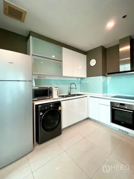 Condominium for rent in Khlong Toei, Bangkok - Modern L-shaped kitchen with oven, microwave, fridge, and washer beneath glossy cabinets.