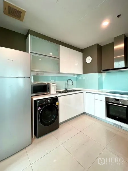 Condominium for rent in Khlong Toei, Bangkok - Modern L-shaped kitchen with oven, microwave, fridge, and washer beneath glossy cabinets.