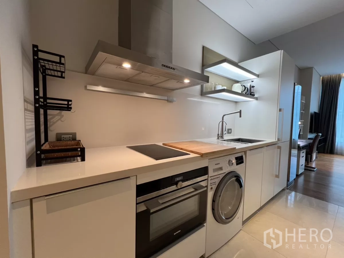 Condominium for rent in Pathum Wan, Bangkok - Modern galley kitchen with induction hob, oven and washer-dryer beneath sleek white countertops.