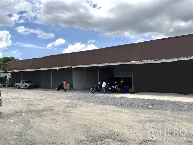 Warehouse for rent in Lam Luk Ka, Pathum Thani - Exterior of single-storey warehouse with multiple roller shutters and a wide gravel parking and loading area under a blue sky in Lam Luk Ka, Pathum Thani.