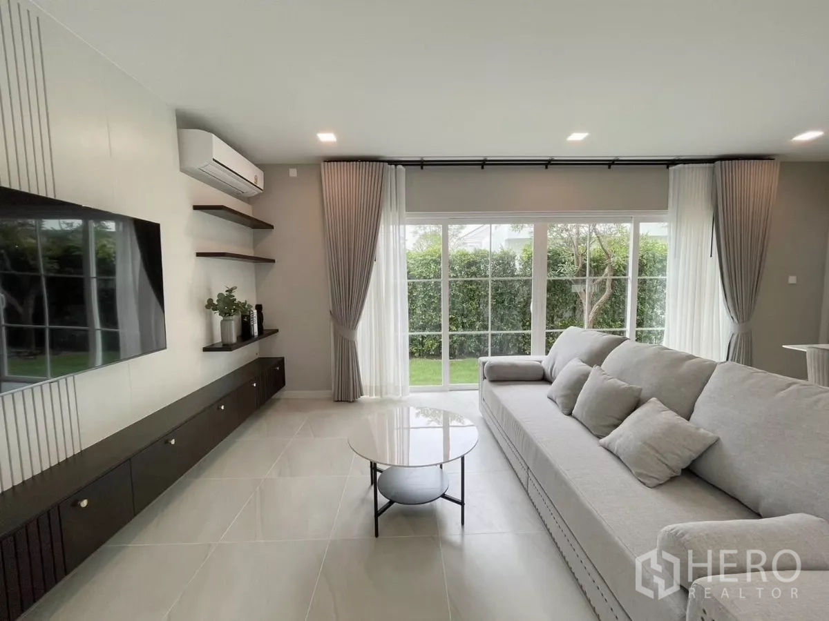 Detached House for rent in Bang Phli, Samut Prakan - Bright living room with large gray sofa, TV wall, and sliding glass doors opening to the garden.