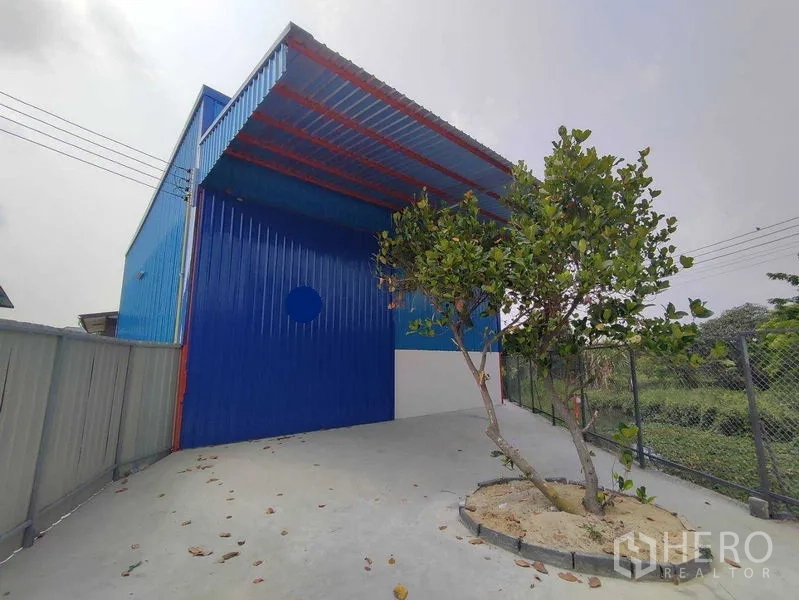 Warehouse for rent in Pak Kret, Nonthaburi - Front driveway and covered loading bay of the blue steel-clad warehouse with side fencing.