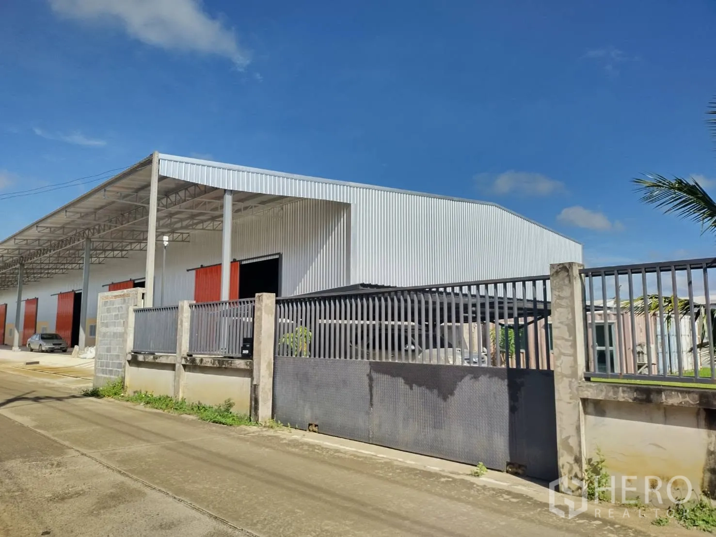 Warehouse for rent in Khlong Luang, Pathum Thani - Corner view of the gated warehouse compound with perimeter fencing and roofed bays along the street.