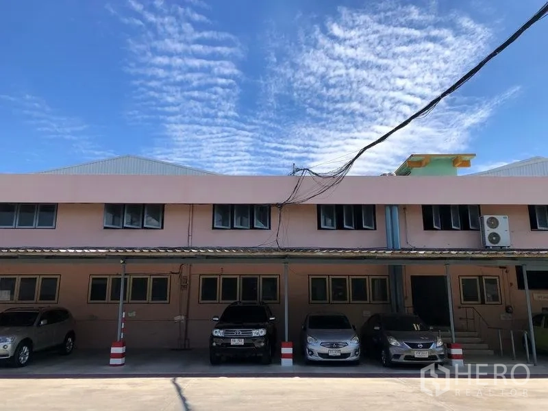 Warehouse for rent in Nonthaburi - Front administration block with covered parking area and several cars under a blue sky.