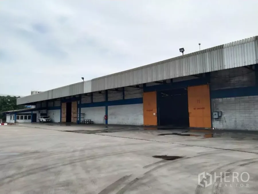 Factory for rent in Samut Prakan - Factory exterior showing wide concrete yard, covered loading bays and large sliding doors with ample truck access in Phra Pradaeng.