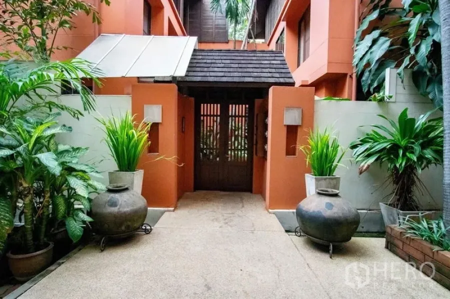 联排别墅 出租在Watthana, Bangkok - 热带风联排别墅入口，赭色外墙、木门与绿植，位于瓦他那区。