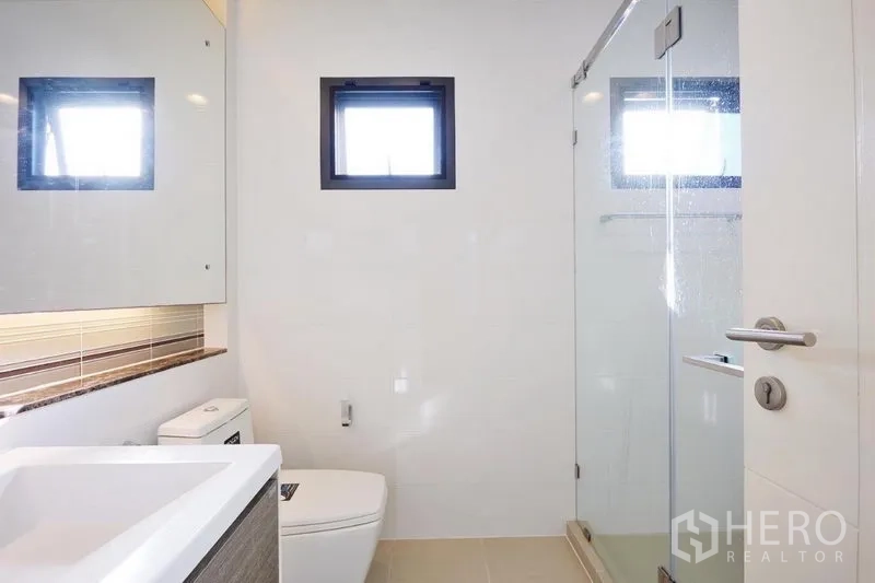 Detached House for rent or sale in Bang Kapi, Bangkok - Minimalist bathroom with glass-enclosed shower, wall-hung vanity and small ventilation windows.