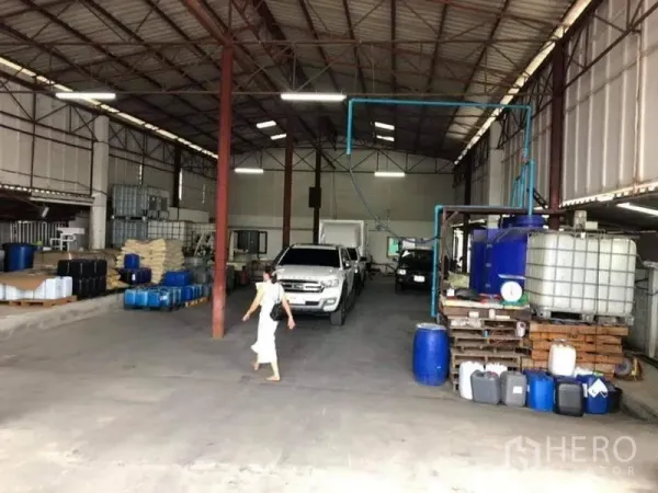 Warehouse for sale in Lam Luk Ka, Pathum Thani - High-roof warehouse bay with pickup trucks, storage drums and containers, bright lighting and a drive-through aisle.