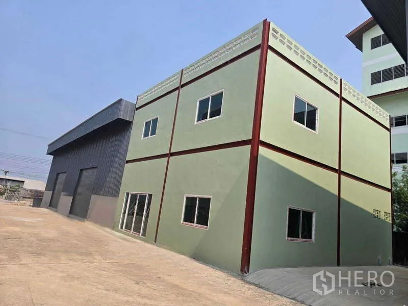 Warehouse for rent in Mueang Samut Sakhon, Samut Sakhon - Two‑storey green office building beside a black steel warehouse and wide concrete yard.