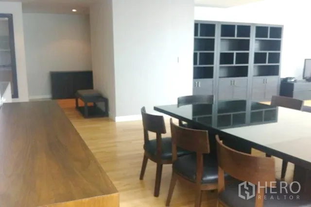 Condominium for sale in Khlong Toei, Bangkok - Open-plan dining area with black glass-top table and wooden chairs beside a long breakfast bar and built-in shelves.