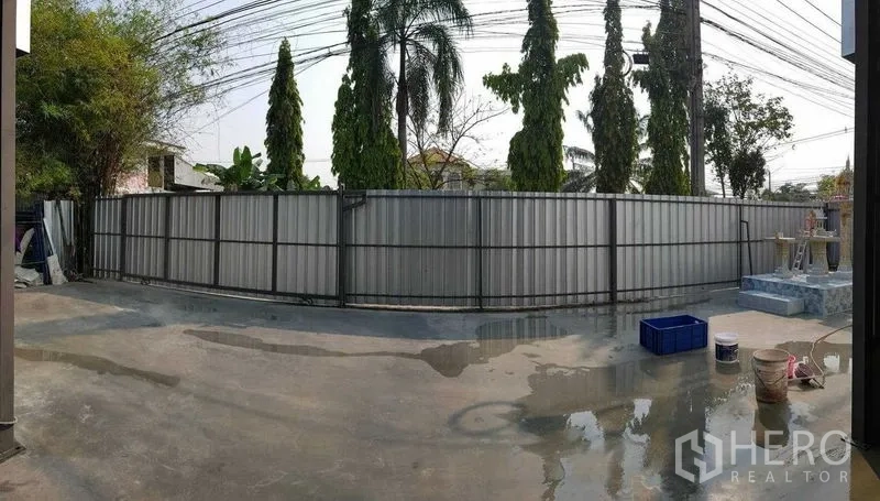 Warehouse for rent in Sai Mai, Bangkok - Perimeter fence of the property with corrugated panels, palm trees outside, and a concrete apron.