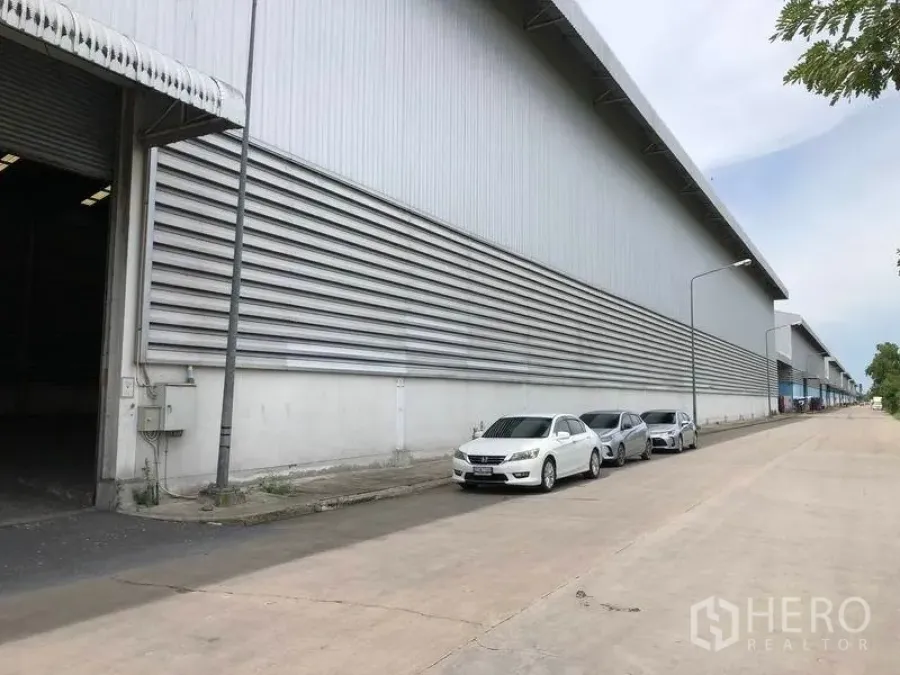 Warehouse for rent in Phra Nakhon Si Ayutthaya - Side exterior of a large metal-clad warehouse with a wide service road and parked cars.