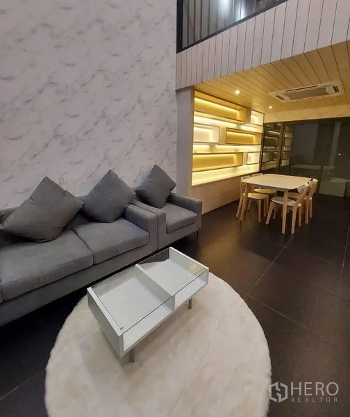 Townhouse for rent in Thanyaburi, Pathum Thani - Open-plan living and dining with gray sofa, illuminated shelving, and wood ceiling.