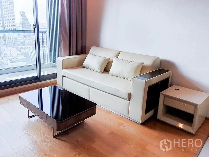 Condominium for rent in Bang Rak, Bangkok - Living room with cream sofa and low coffee table beside a balcony door overlooking the city.