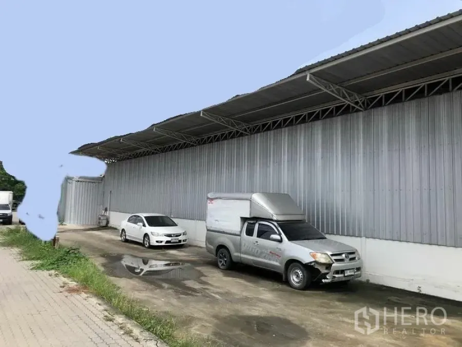 Warehouse for rent in Pak Kret, Nonthaburi - Long metal-clad warehouse exterior with covered loading bay and parked vehicles in Pak Kret, Nonthaburi.