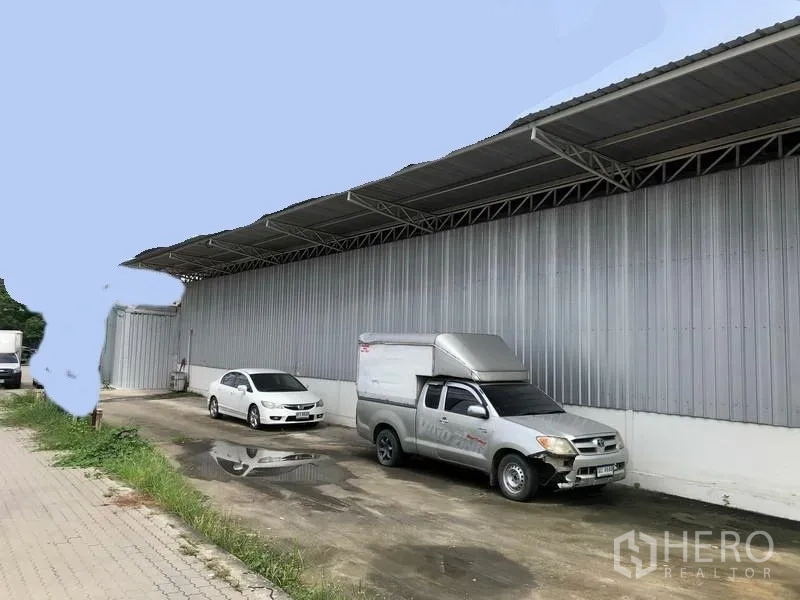 Warehouse for rent in Pak Kret, Nonthaburi - Long metal-clad warehouse exterior with covered loading bay and parked vehicles in Pak Kret, Nonthaburi.