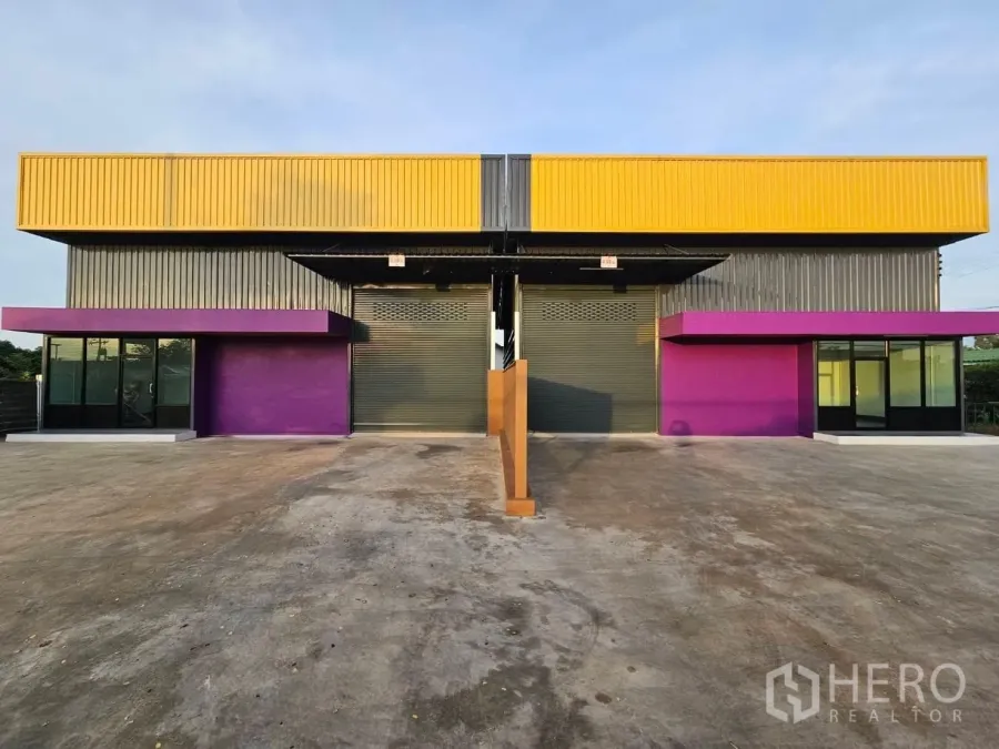 Warehouse for rent in Lam Luk Ka, Pathum Thani - Front view of a modern twin-bay warehouse with yellow cladding, purple canopies, two roller doors and a wide concrete yard in Lam Luk Ka.