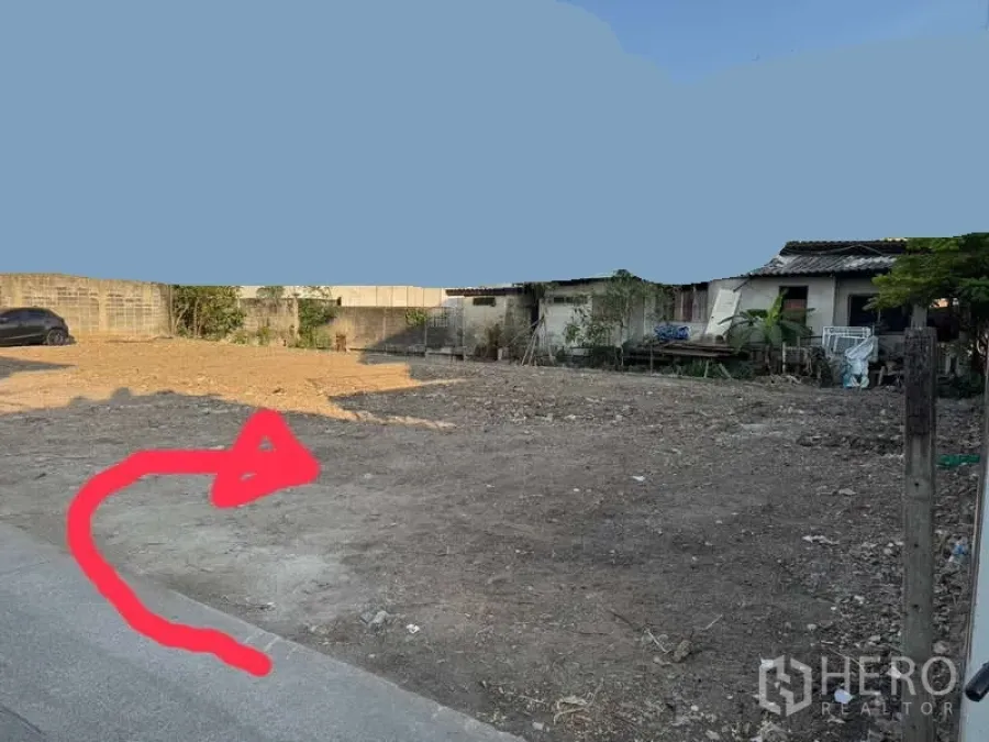 Land for rent in Suan Luang, Bangkok - Cleared vacant land plot with dirt surface, boundary walls, small houses at the edge, and paved road access in Suan Luang, Bangkok, with a red arrow marking the entry.