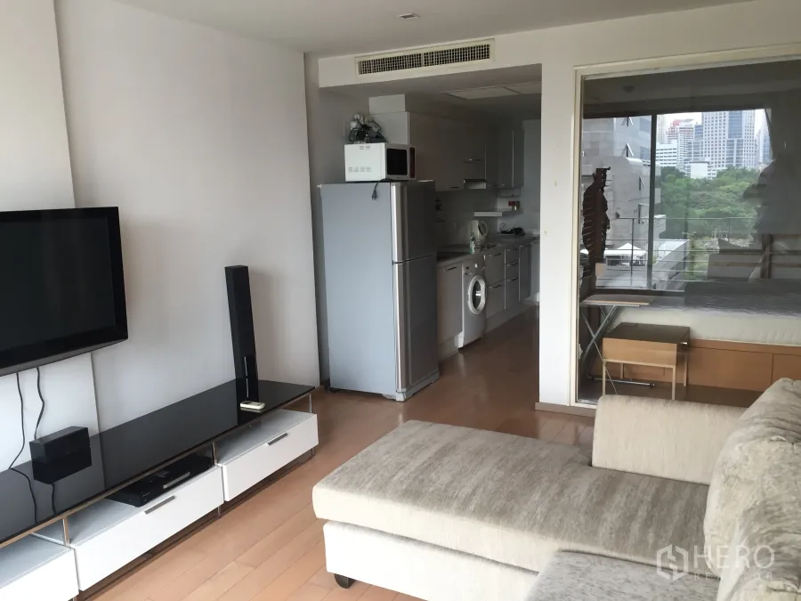 Condominium for rent in Pathum Wan, Bangkok - Open-plan living room with sofa, wall-mounted TV and a fitted kitchen with fridge, microwave and washing machine, plus a glass partition to the bedroom and park-city view.