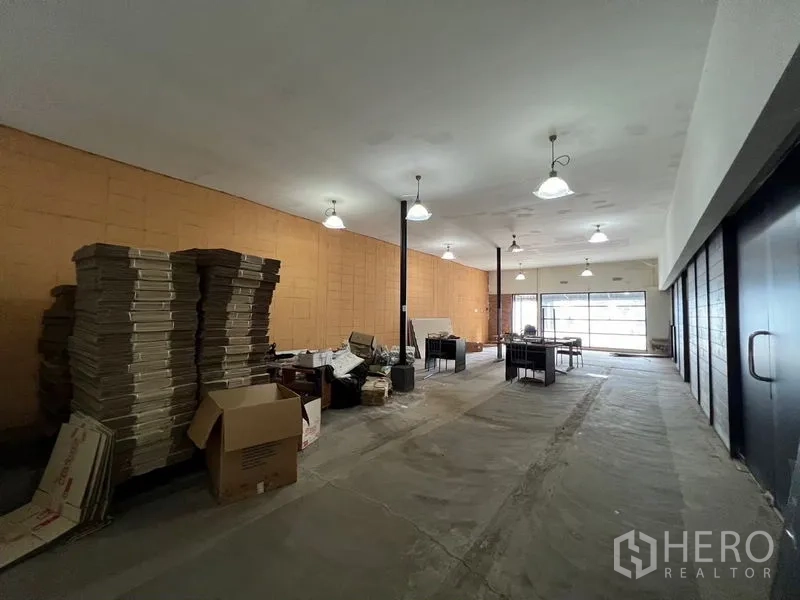Warehouse for rent in Suan Luang, Bangkok - Open-plan warehouse interior featuring high ceilings, pendant lights, concrete flooring, and front glazing with stacked cartons.