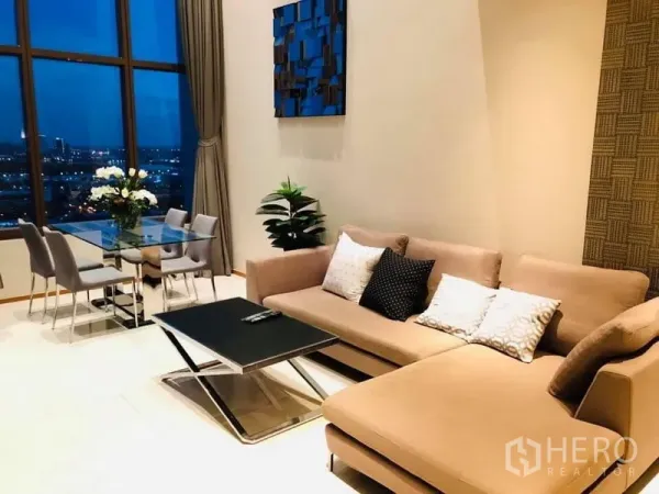 Condominium for rent in Khlong Toei, Bangkok - Cozy living room with L-shaped sofa and glass dining table facing city lights in a Bangkok condo.