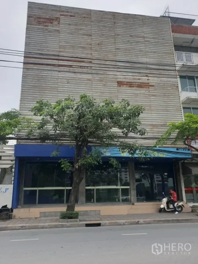 Retail Space for rent in Watthana, Bangkok - Street-facing retail building with a full-width glass storefront, blue awning, and mature tree along the sidewalk.