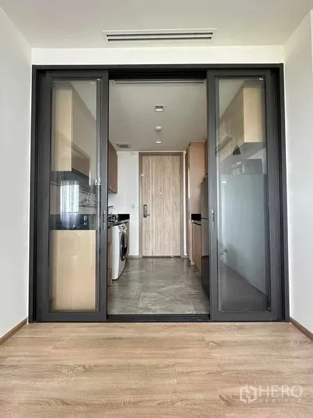Condominium for sale in Watthana, Bangkok - Living room with wood floors facing an enclosed kitchen through black‑framed sliding glass doors.