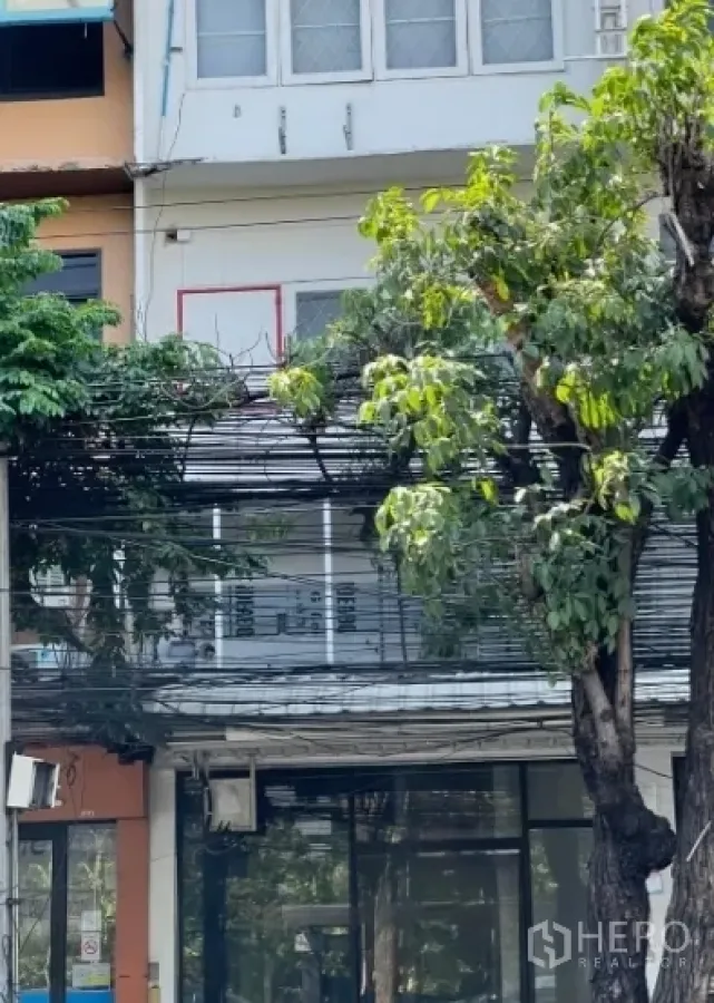 Shophouse for sale in Watthana, Bangkok - Ground-floor shopfront with large display windows beneath mature trees, part of an urban shophouse row near BTS Phra Khanong.