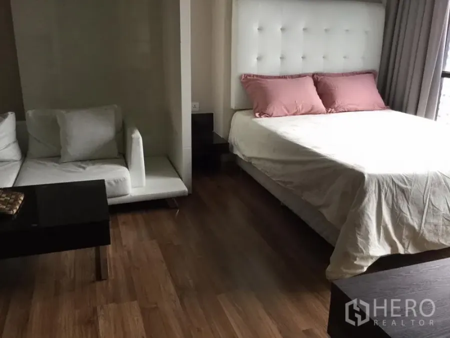 Condominium for rent in Huai Khwang, Bangkok - Studio-style bedroom with padded headboard, white sofa nook and wood flooring beside a large window.