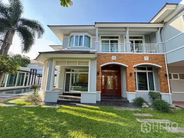 Detached House for rent or sale in Prawet, Bangkok - Front facade with arched brick accents, upper balcony and lush green lawn.