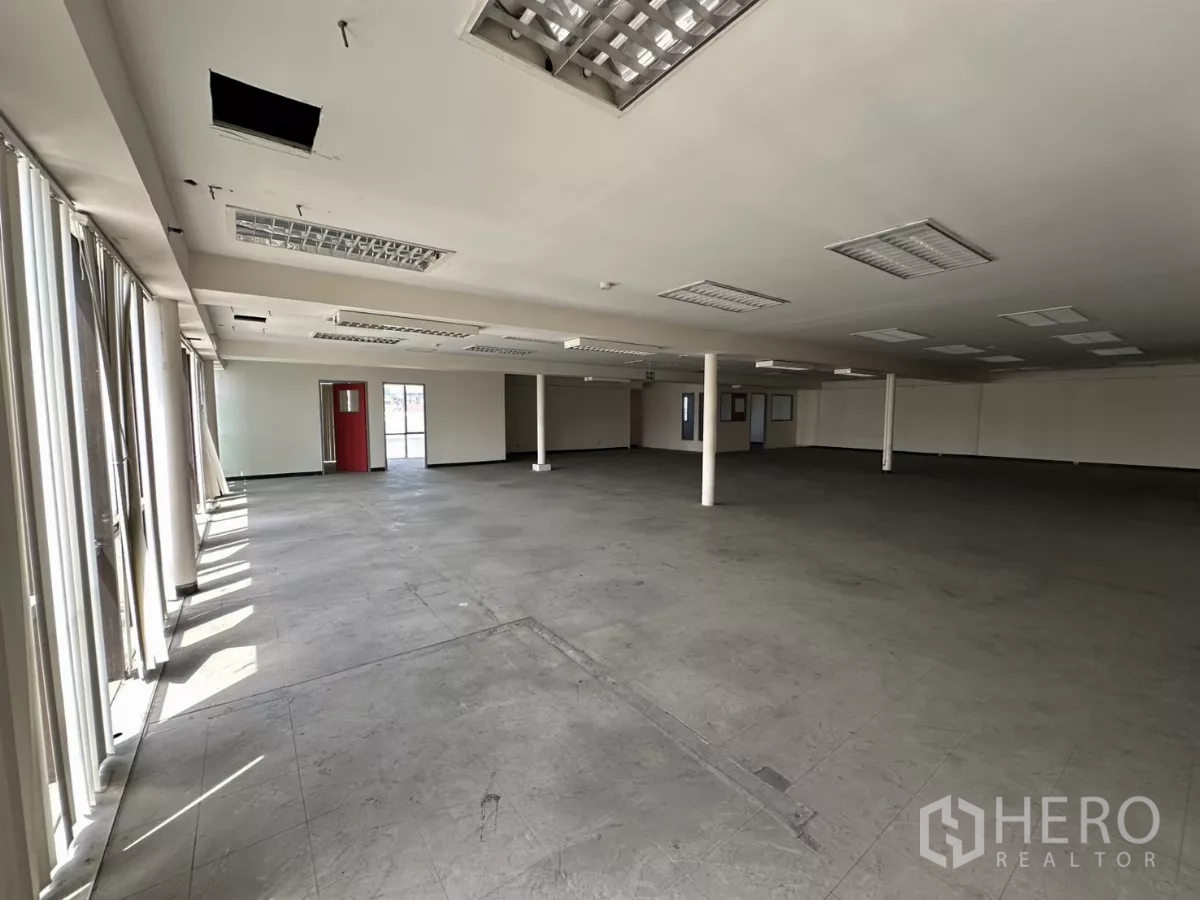 Warehouse for rent in Suan Luang, Bangkok - Expansive office floor with columns, a wall of windows with vertical blinds, and tiled ceiling panels.