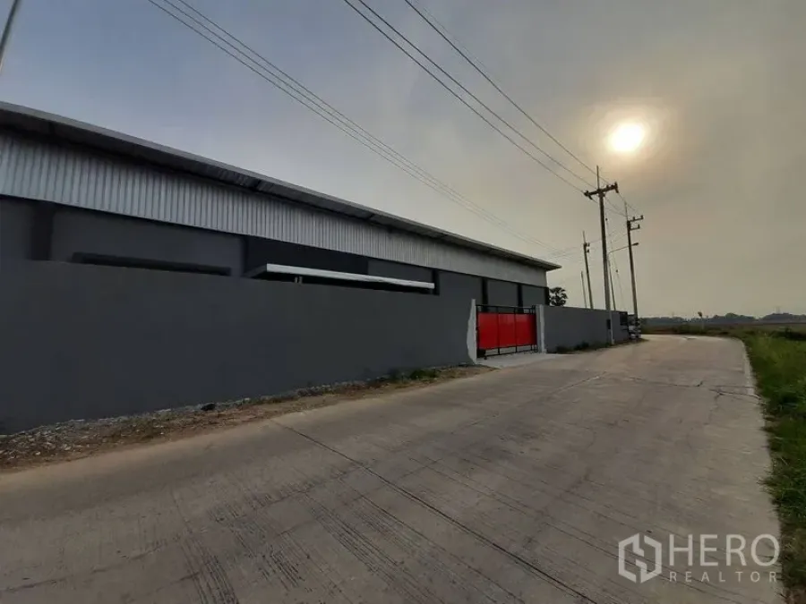Warehouse for rent in Lam Luk Ka, Pathum Thani - Street view of the gated warehouse wall and red entrance gate along a concrete road.