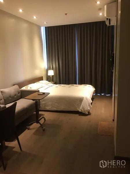 Condominium for rent in Khlong Toei, Bangkok - Cozy studio-style bedroom with double bed, grey blackout curtains, sofa and side table under recessed ceiling lights.