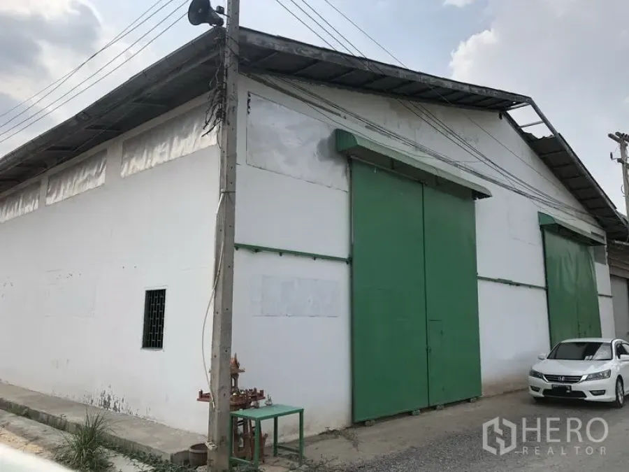 Warehouse for rent or sale in Khlong Luang, Pathum Thani - Street-side view of a white warehouse with large green sliding doors and a metal roof in Khlong Luang, Pathum Thani.