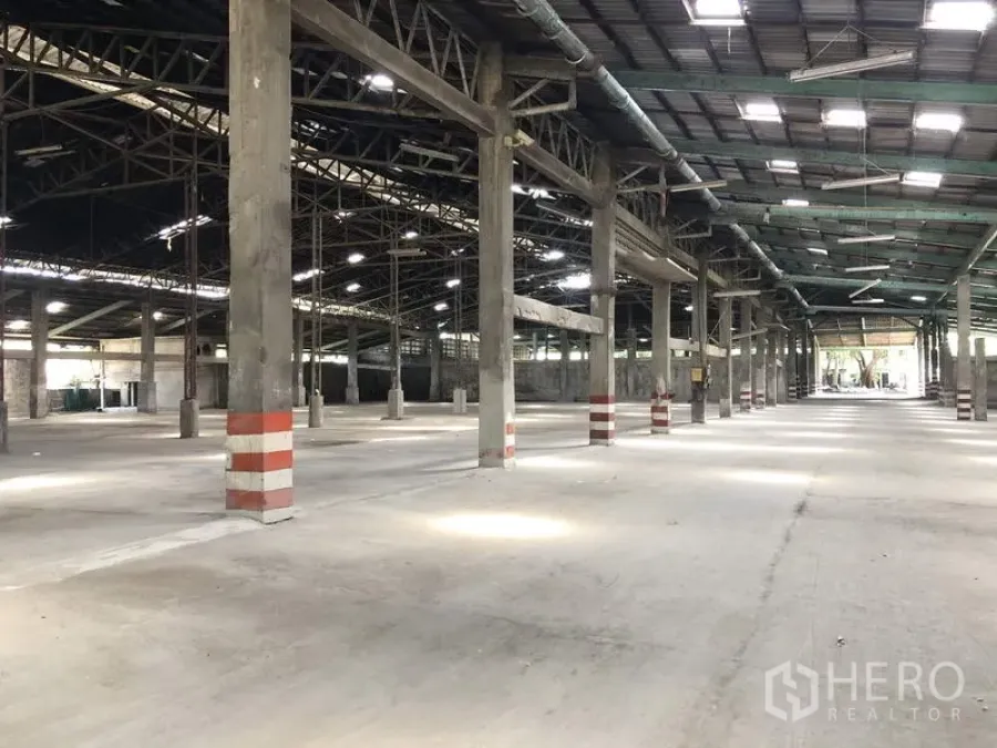 Warehouse for rent in Thanyaburi, Pathum Thani - Vast warehouse interior with concrete floor, high steel-truss roof, skylights and striped safety columns.