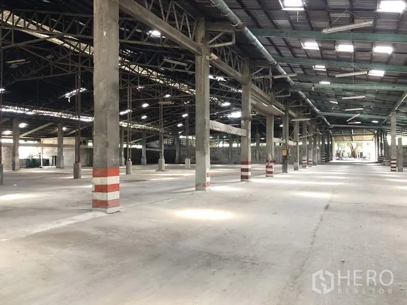 Warehouse for rent in Thanyaburi, Pathum Thani - Vast warehouse interior with concrete floor, high steel-truss roof, skylights and striped safety columns.