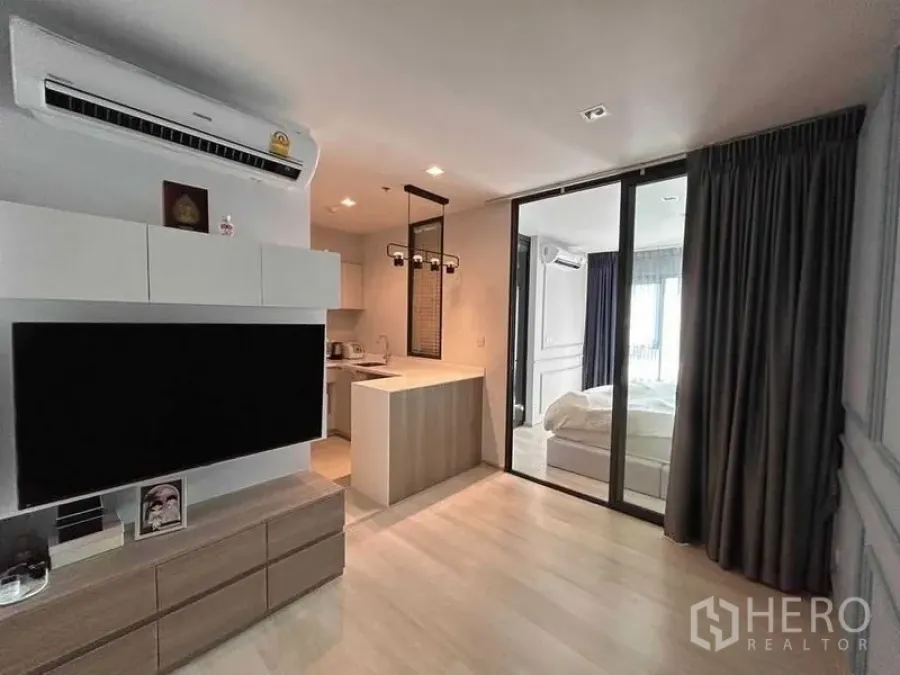 Condominium for sale in Pathum Wan, Bangkok - Open‑plan living area with TV console, kitchenette and breakfast bar, sliding glass doors to the bedroom, and blackout drapes.