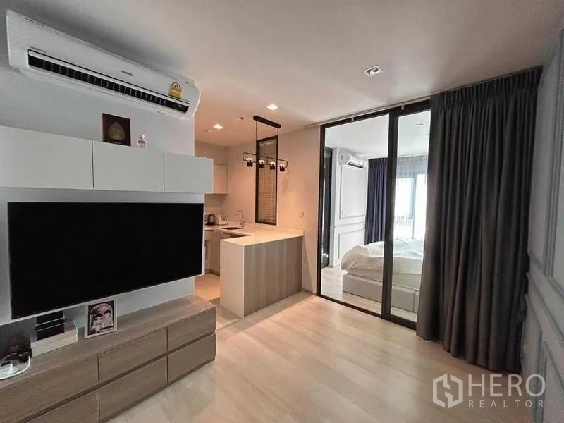 Condominium for sale in Pathum Wan, Bangkok - Open‑plan living area with TV console, kitchenette and breakfast bar, sliding glass doors to the bedroom, and blackout drapes.