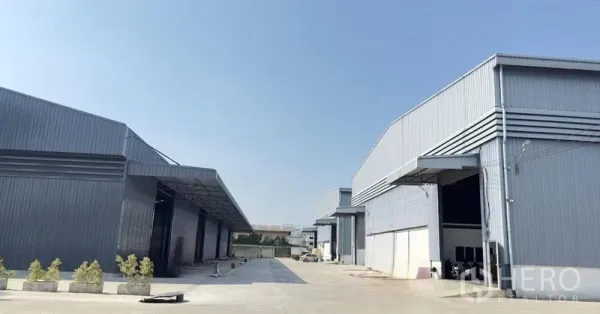 Warehouse for rent in Pathum Thani - Modern steel-clad warehouse buildings with wide concrete driveway and covered loading bays under a clear sky.