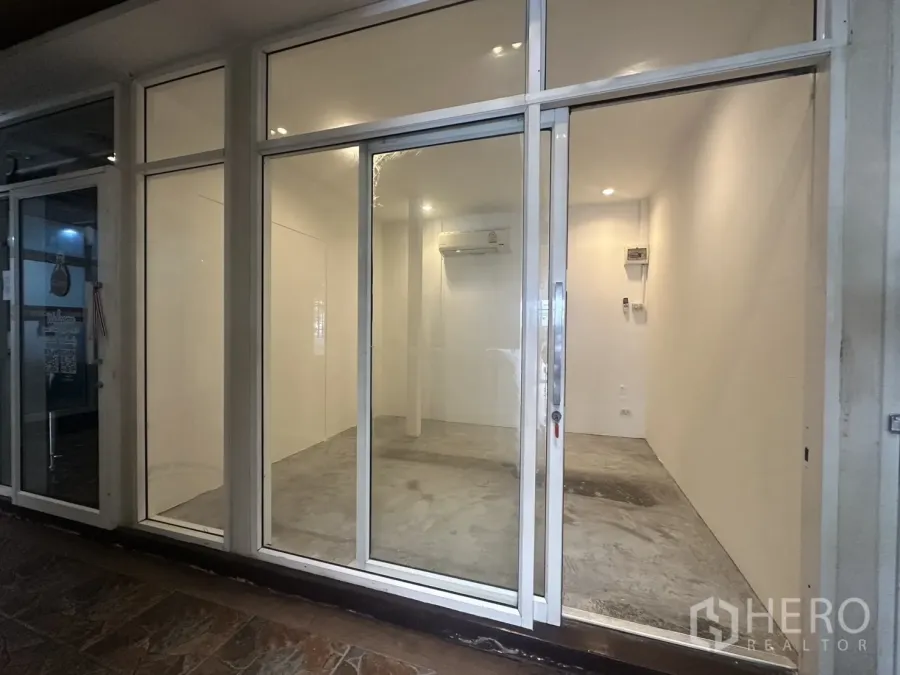 Retail Space for rent - Glass-fronted retail unit with sliding door, white walls, concrete floor, recessed lights and air conditioner in a boutique mall corridor near BTS Phrom Phong.