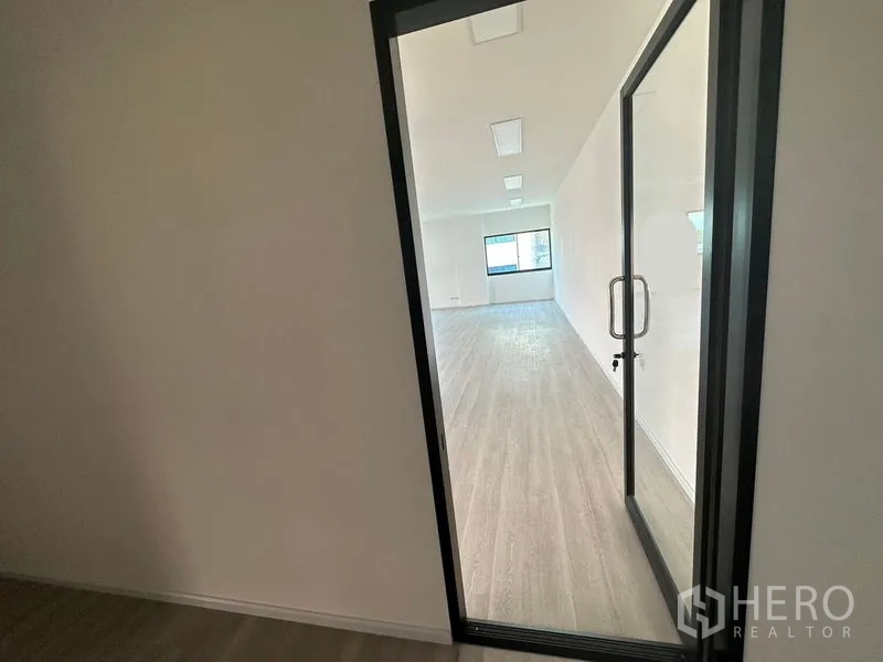 Office Space for rent - View through a glass door into a bright open-plan office with wood-look flooring, white walls, ceiling lights and a window at the far end.